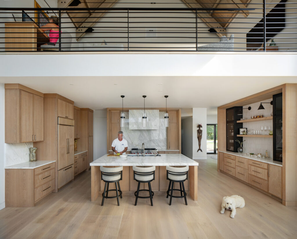 A man cuts fruit in his neutral, open concept kitchen with his golden retriever near by.
