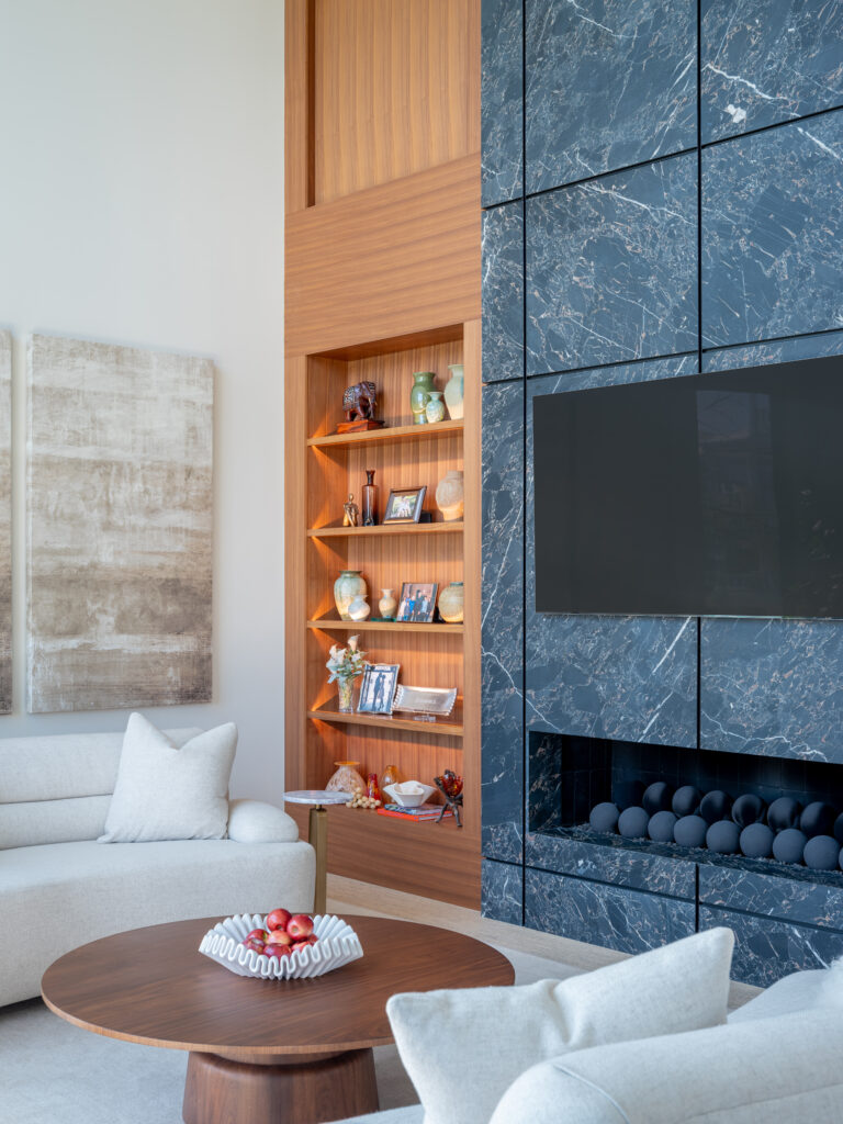 A modern and coastal living room with plush rounded couches, a circular fireplace, walnut cabinetry, and a floor to ceiling dekton fireplace that is flanked by walnut cabinets.