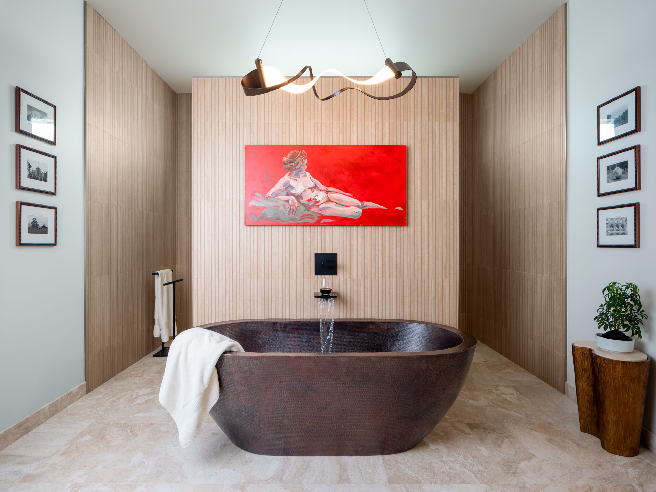 A master bathroom is pictured with a large, copper soaking tub at the center underneath a striking red painting. Gallery walls flank either side of the tub in the center. The tub is set against the opposite side of the zero-entrance shower wall.