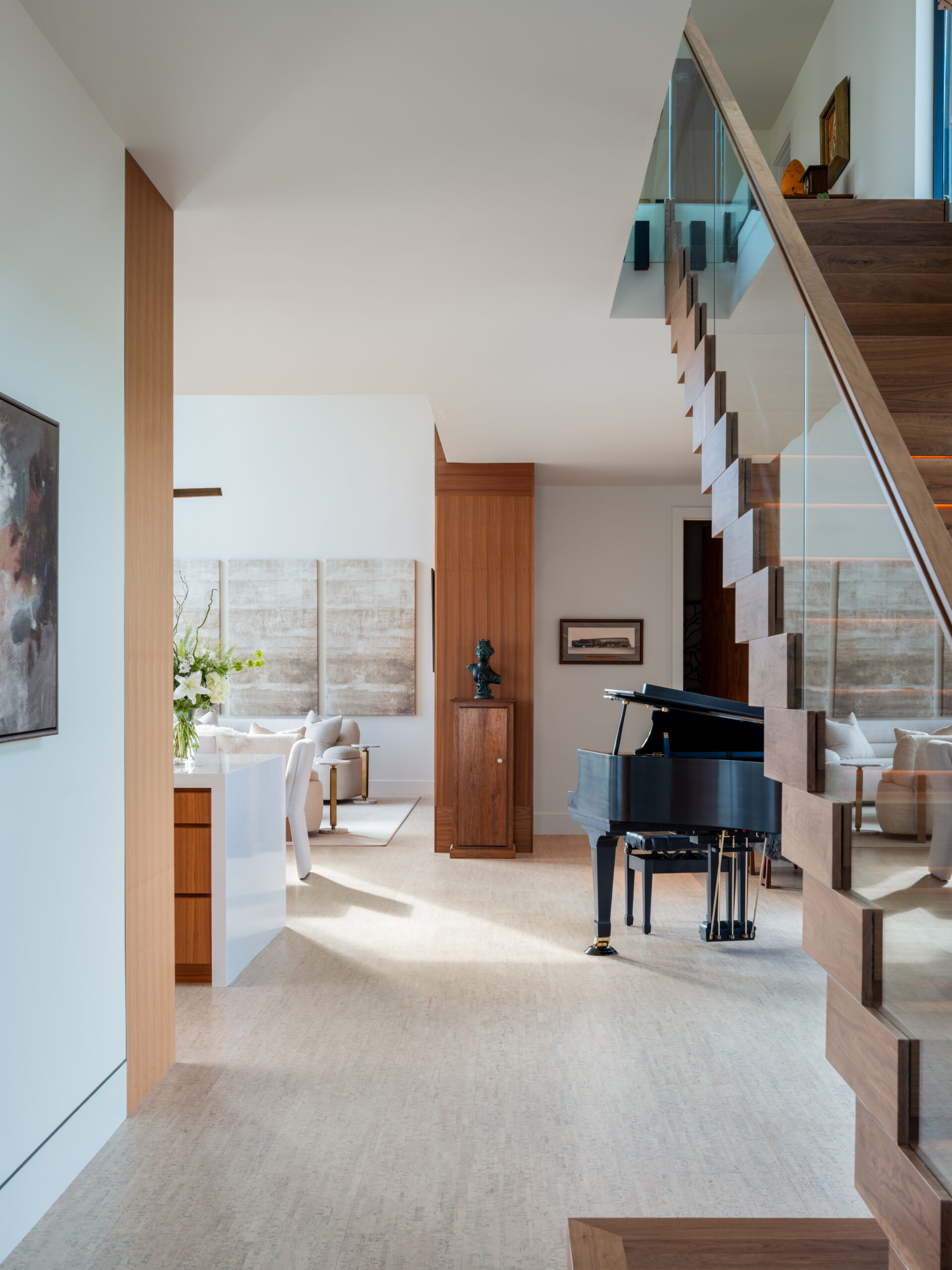 A shot of the sightline through the main living space and heart of this home. We can see a modern floating staircase, behind it a piano, and across is the kitchen.