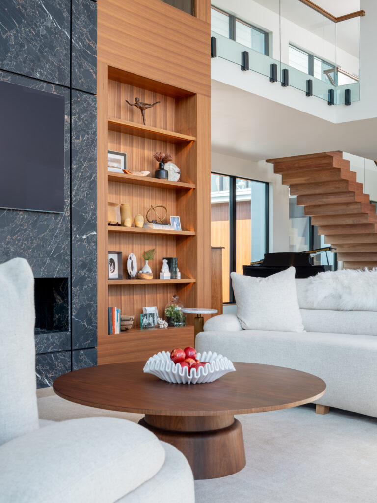 A modern and coastal living room with plush rounded couches, a circular fireplace, walnut cabinetry, and a floor to ceiling dekton fireplace that is flanked by walnut cabinets.