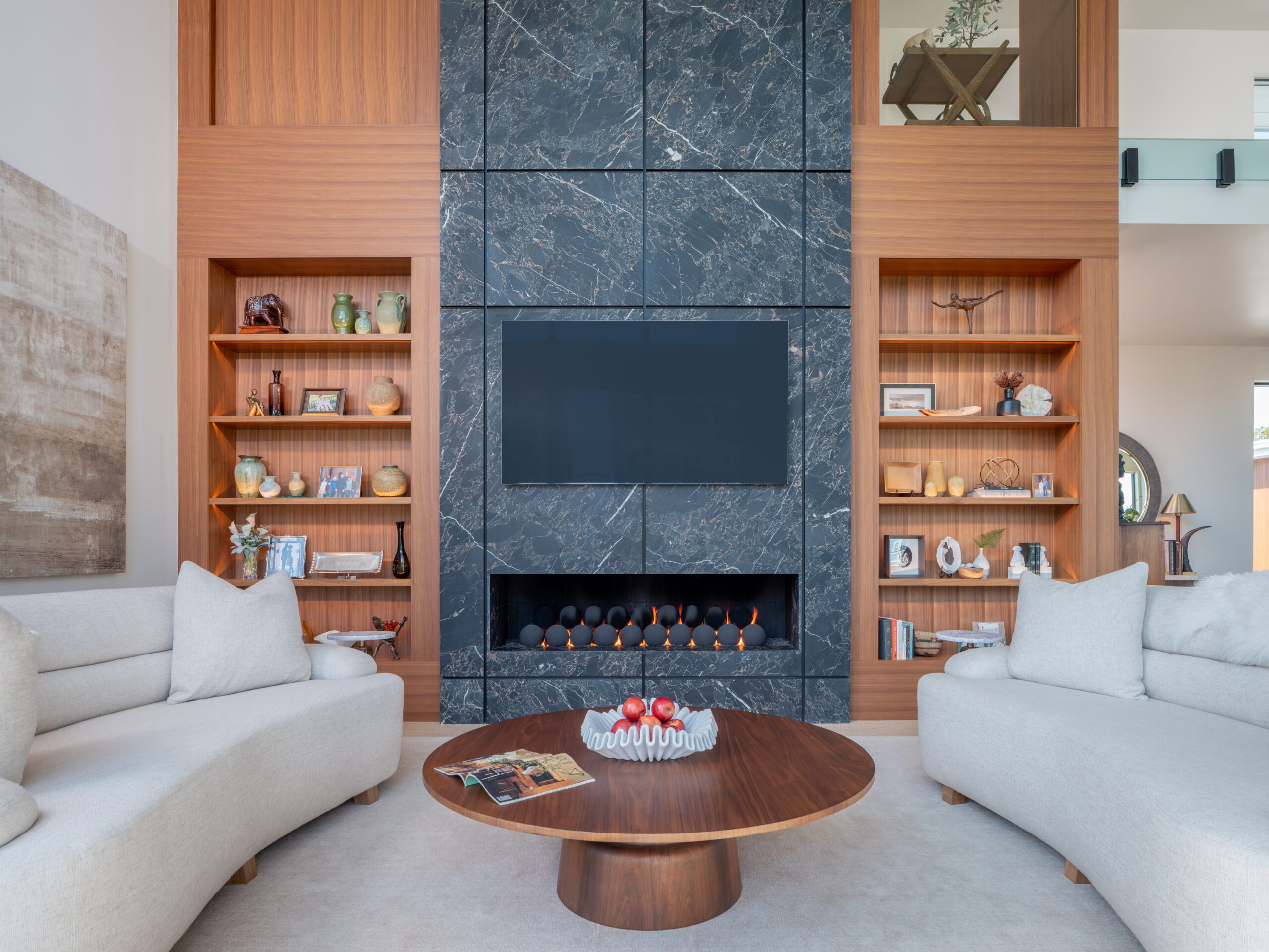 A modern and coastal living room with plush rounded couches, a circular fireplace, walnut cabinetry, and a floor to ceiling dekton fireplace that is flanked by walnut cabinets.